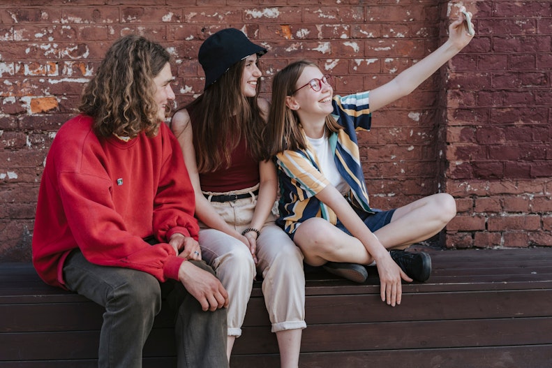 Three teenagers sit together on a bench in front of a brick wall, laughing while one takes a selfie.