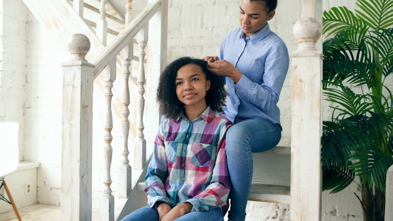 A mother sits on a staircase styling her daughter's curly hair while the daughter sits on a lower step.