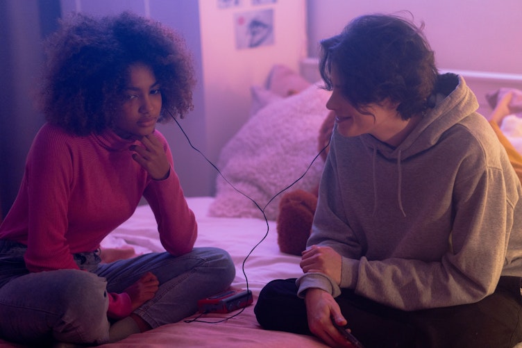 Two teenagers sit on a bed sharing a pair of wired earphones, lit in purple and pink tones.