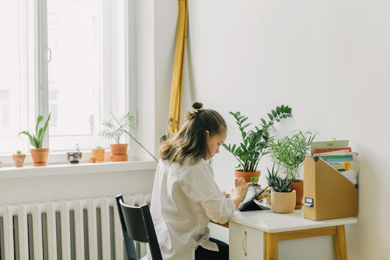 A woman sits at a small desk by a window surrounded by potted plants, working on a tablet.