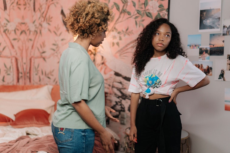 A woman and a teenage girl stand facing each other in a bedroom, appearing to be in a tense conversation.