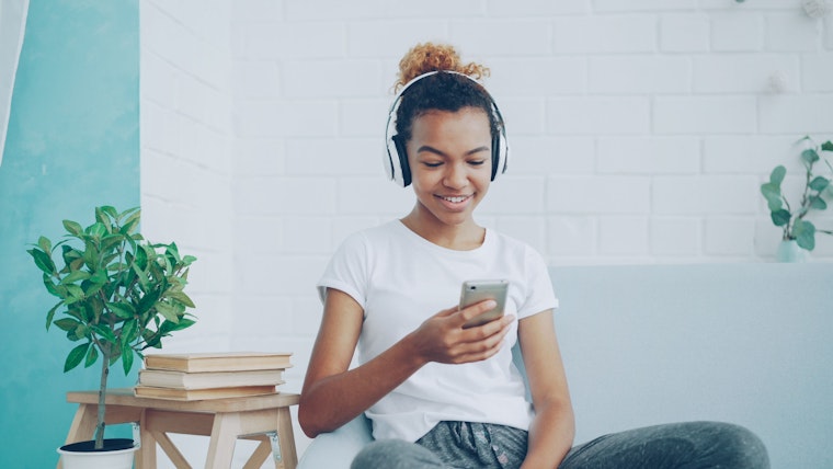A teenage girl wears headphones and smiles while looking at her phone, sitting on a couch near a plant and a stack of books.