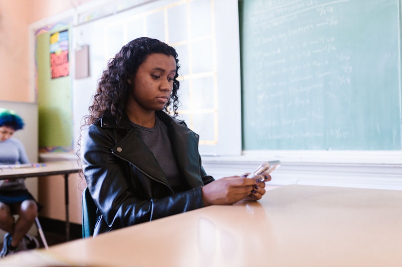 A teenage girl sits at a desk in a classroom looking down at her phone, with a chalkboard and another student in the background.