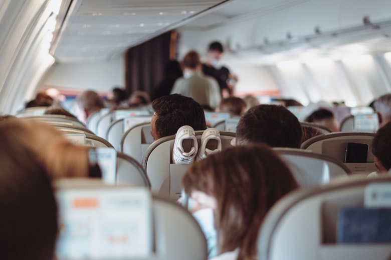 Passengers sit in aeroplane seats with shoes propped up on a headrest, while people stand in the aisle near the front of the cabin.