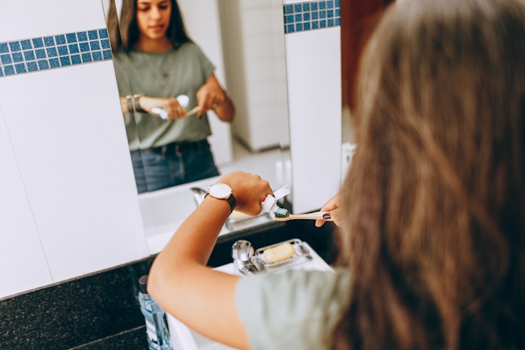 A girl applies toothpaste to a bamboo toothbrush while facing a bathroom mirror.