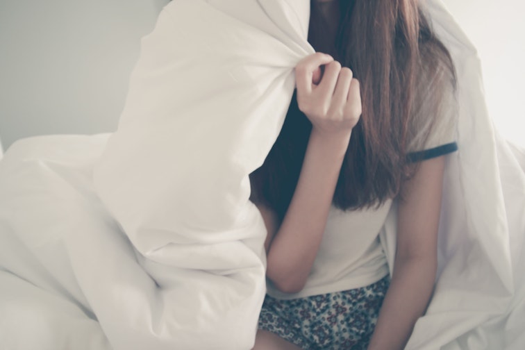 A girl sits on a bed with a white sheet pulled up and over most of her head and body, hiding underneath it.