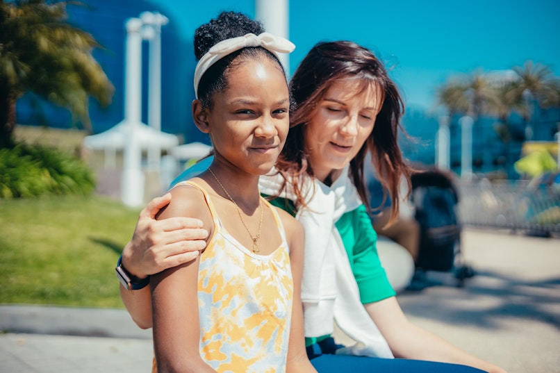 A woman rests her hand on a teenage girl's shoulder as they sit together outdoors, both facing the camera.