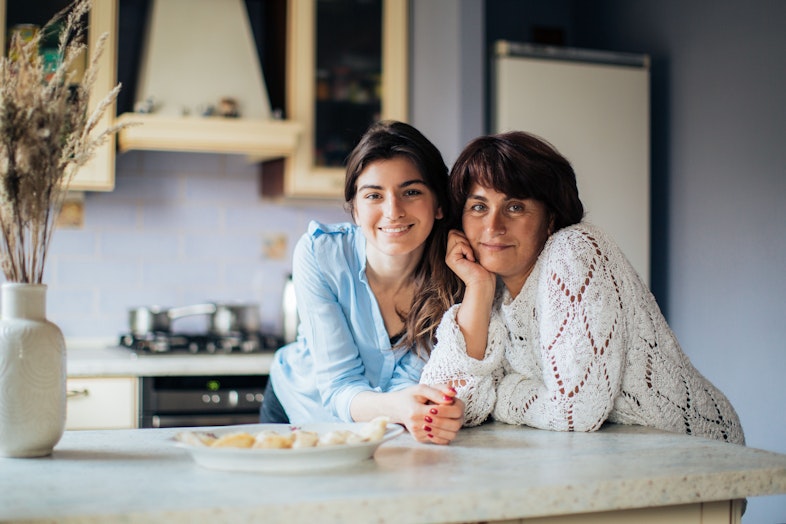 A mother and daughter lean together on a kitchen counter smiling, with a plate of food in front of them.