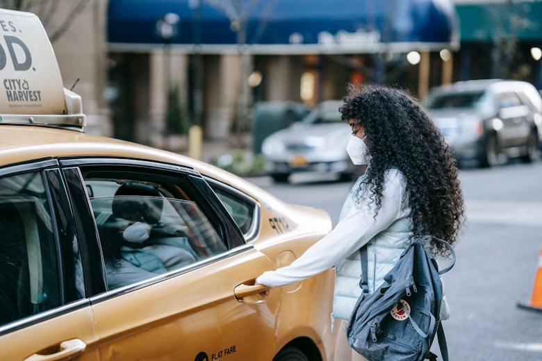 A teenage girl wearing a face mask and carrying a backpack opens the door of a yellow taxi on a city street.