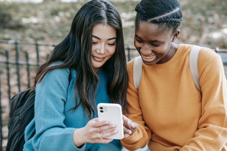 Two teenage girls with backpacks stand outdoors smiling at a phone one of them is holding.