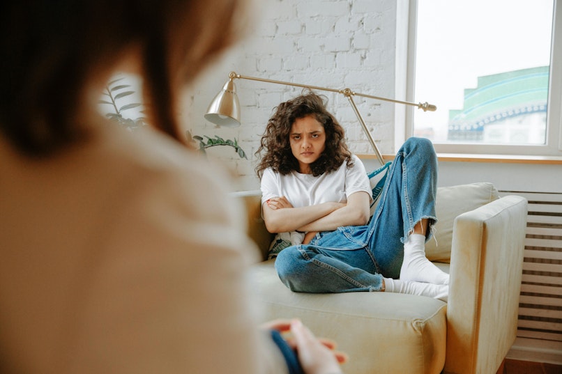 A teenage girl sits on a sofa with her arms crossed and a frustrated expression, facing another person seen from behind.