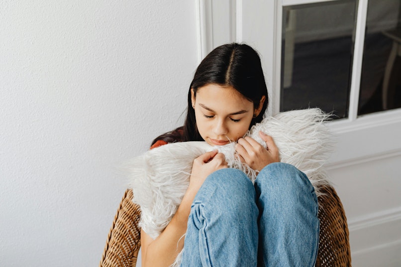 A girl sits curled up in a wicker chair hugging a fluffy white cushion, eyes closed.