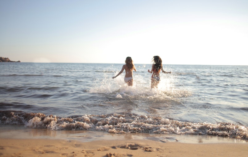 Two teenage girls in swimwear running through the sea at the beach, splashing water around them.