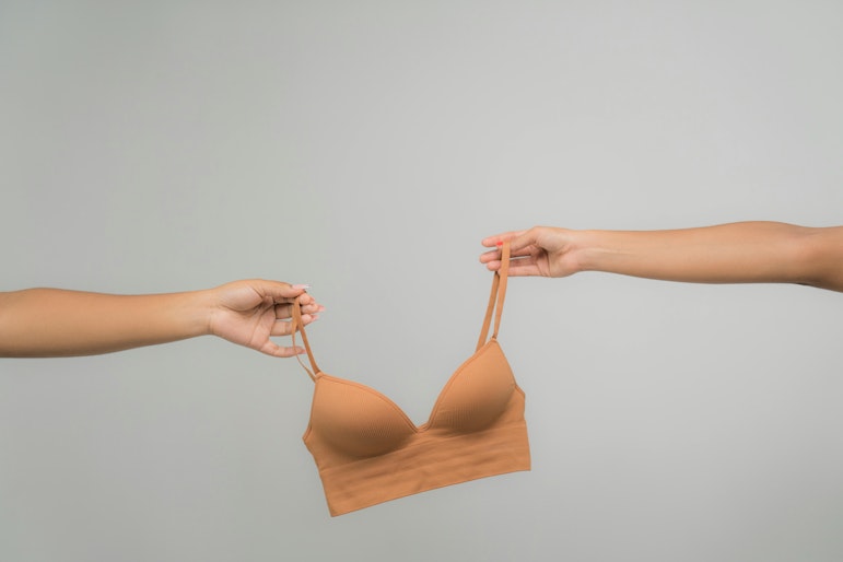 Two hands holding either strap of an orange bralette, passing it between them against a plain grey background.