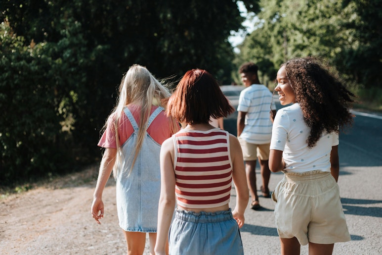 A group of teenagers walking together along a road lined with trees, seen from behind.