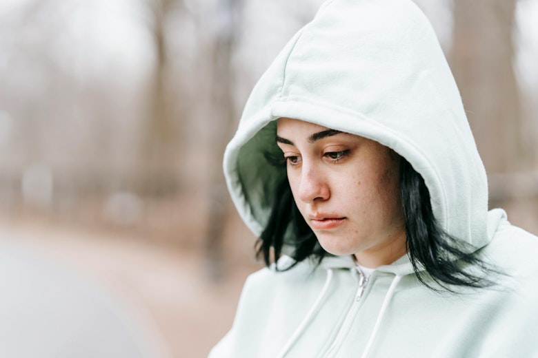 Teenage girl in a hooded jacket looking down, standing outdoors in a wooded area.