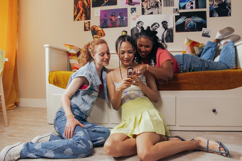Three teenage girls sitting on the floor together looking at a phone, in a bedroom decorated with posters.
