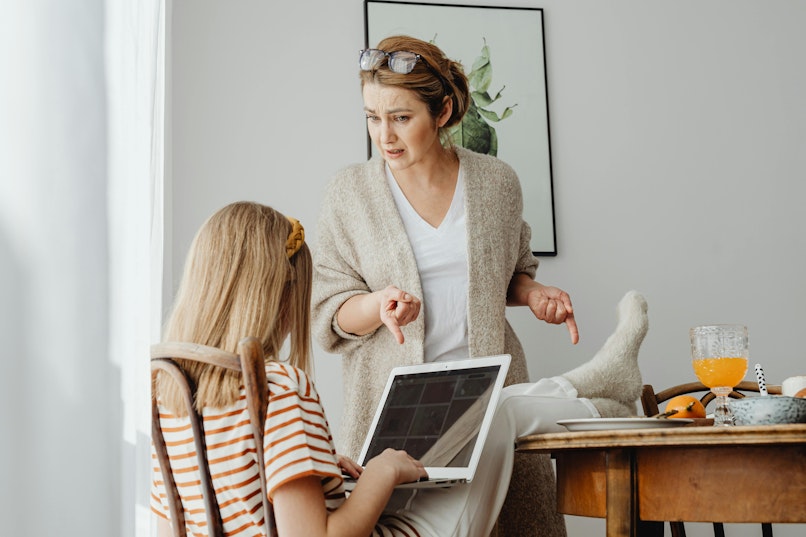 A mother standing and gesturing while pointing at her tween daughter, who is sitting at the table with a laptop.