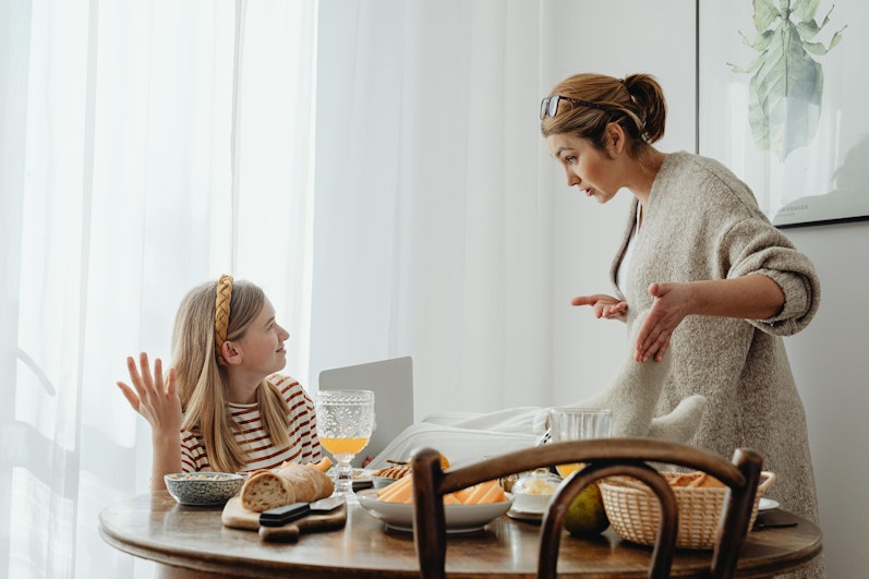 A mother stands talking with her daughter, who is seated at a breakfast table, in a bright room.