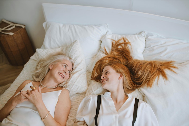 A mother and daughter lie on a bed smiling at each other, with the daughter's red hair fanned out on the pillow.