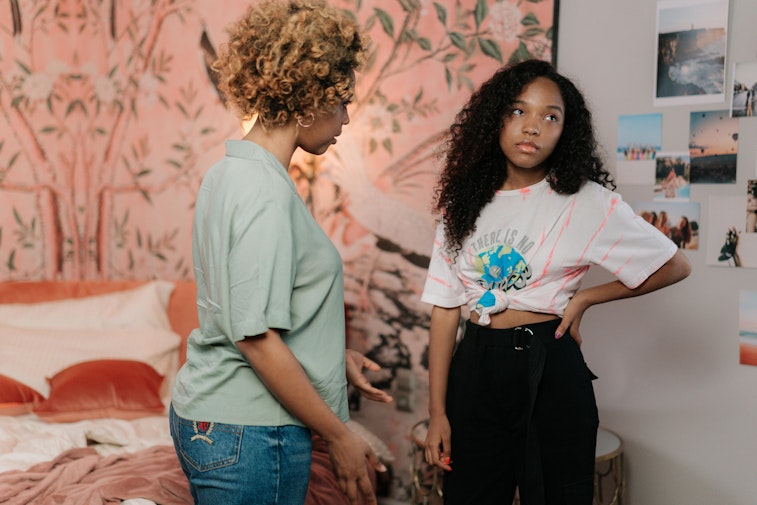 A woman talks with a teenage girl standing with her hand on her hip in a bedroom decorated with photos and floral wallpaper.