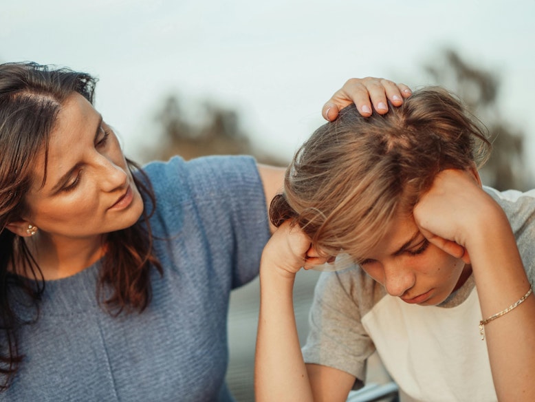A mother comforts her son, who looks upset with his head resting on his hand, placing a hand on his head outdoors.