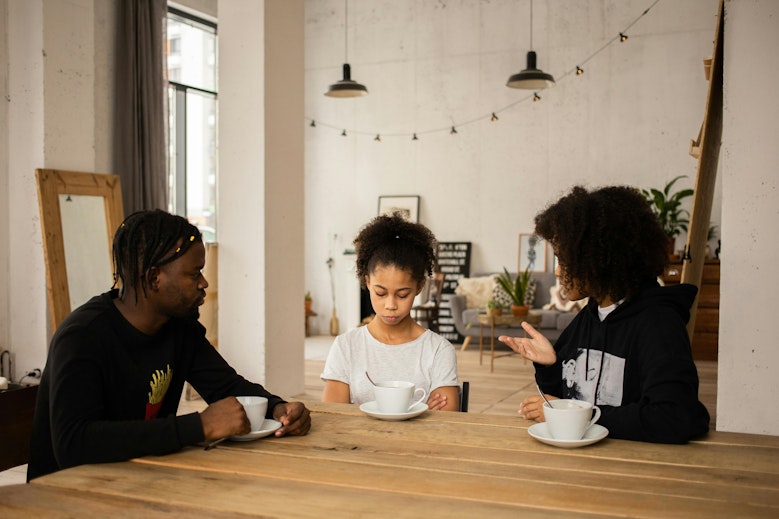 A man and woman sit at a table with a teenage girl who looks down at her cup, having a serious conversation over coffee.