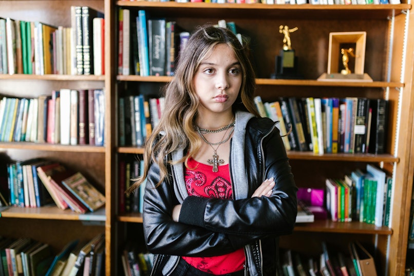 A teenage girl in a leather jacket stands with her arms crossed and a serious expression in front of bookshelves.