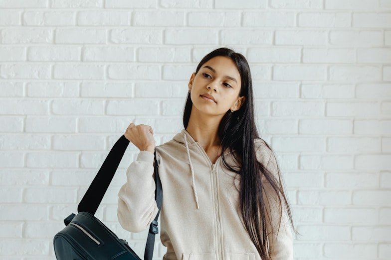 A teenage girl with long dark hair carries a bag over her shoulder against a white brick wall.