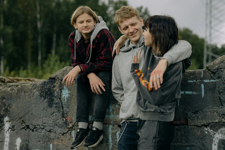 Three teenagers sit and stand together on a low concrete wall outdoors, with one boy putting his arm around a girl.