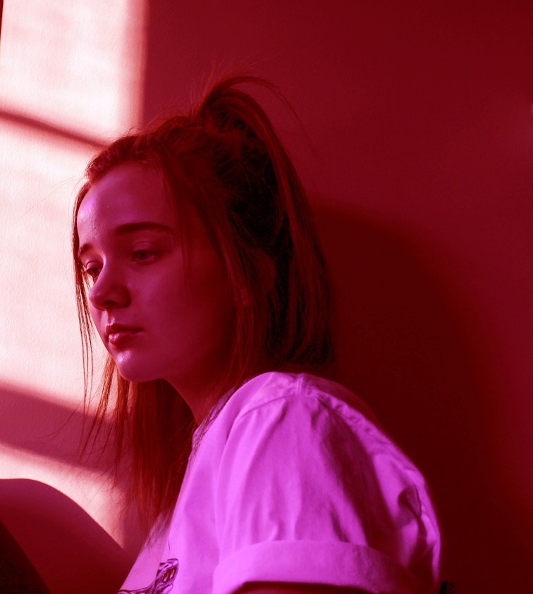 A teenage girl with her hair in a bun sits looking down, lit by pink and red tones.