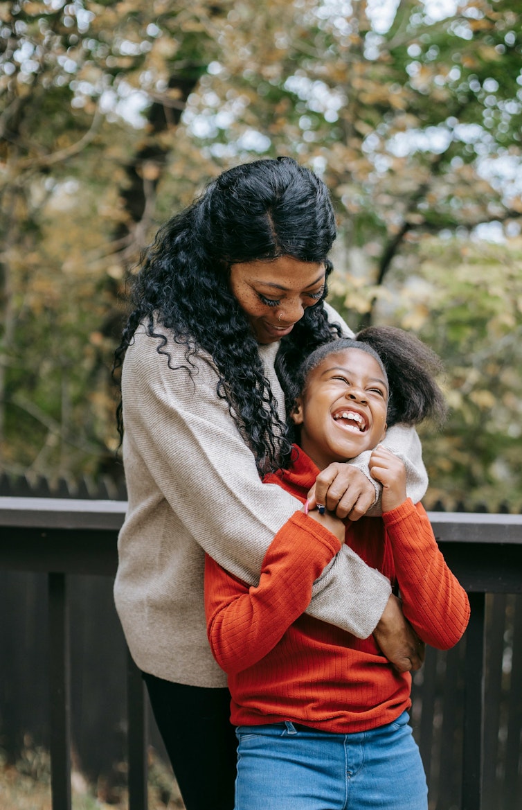 A mother holds and laughs with her young daughter outdoors, both smiling.