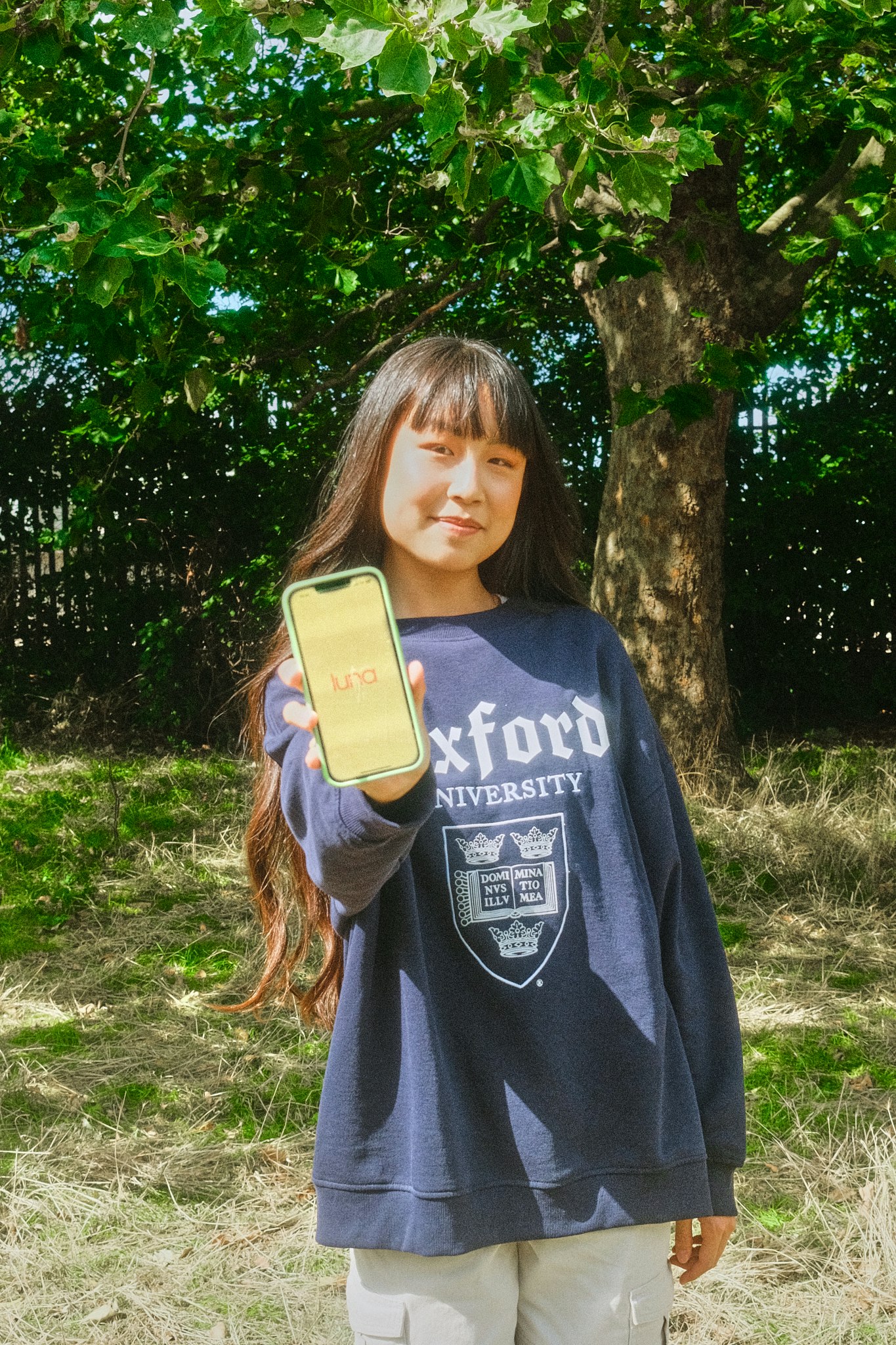 Teenage girl with long hair, wearing an Oxford University sweatshirt, stands outdoors among trees holding up a phone showing the luna logo on screen.