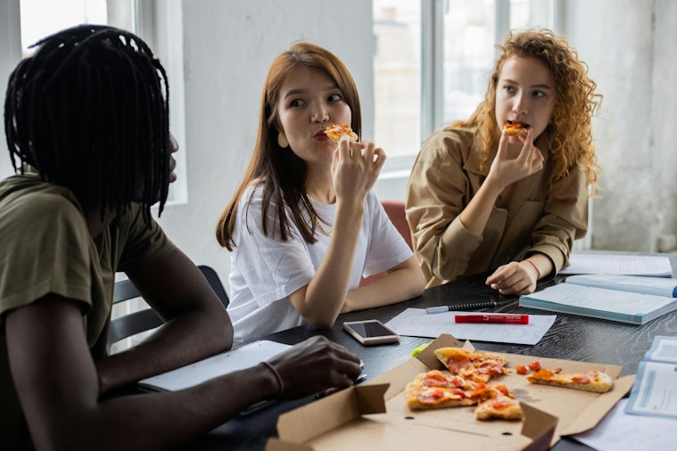Three teenage girls sitting at a table eating pizza together, with notebooks, a pen and a phone on the table.