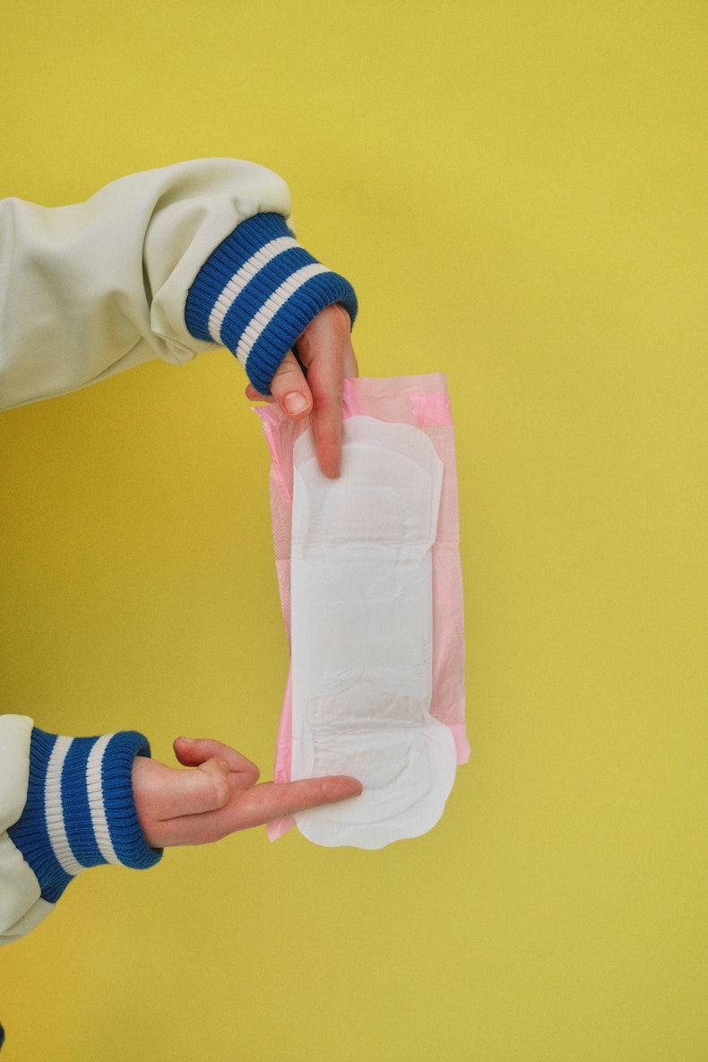 Two hands wearing a striped cuffed jumper unfold a disposable period pad against a yellow background.