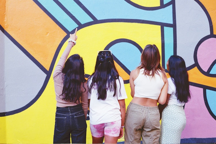 Four teenage girls seen from behind, standing in front of a colourful graffiti-style mural, one pointing upward.