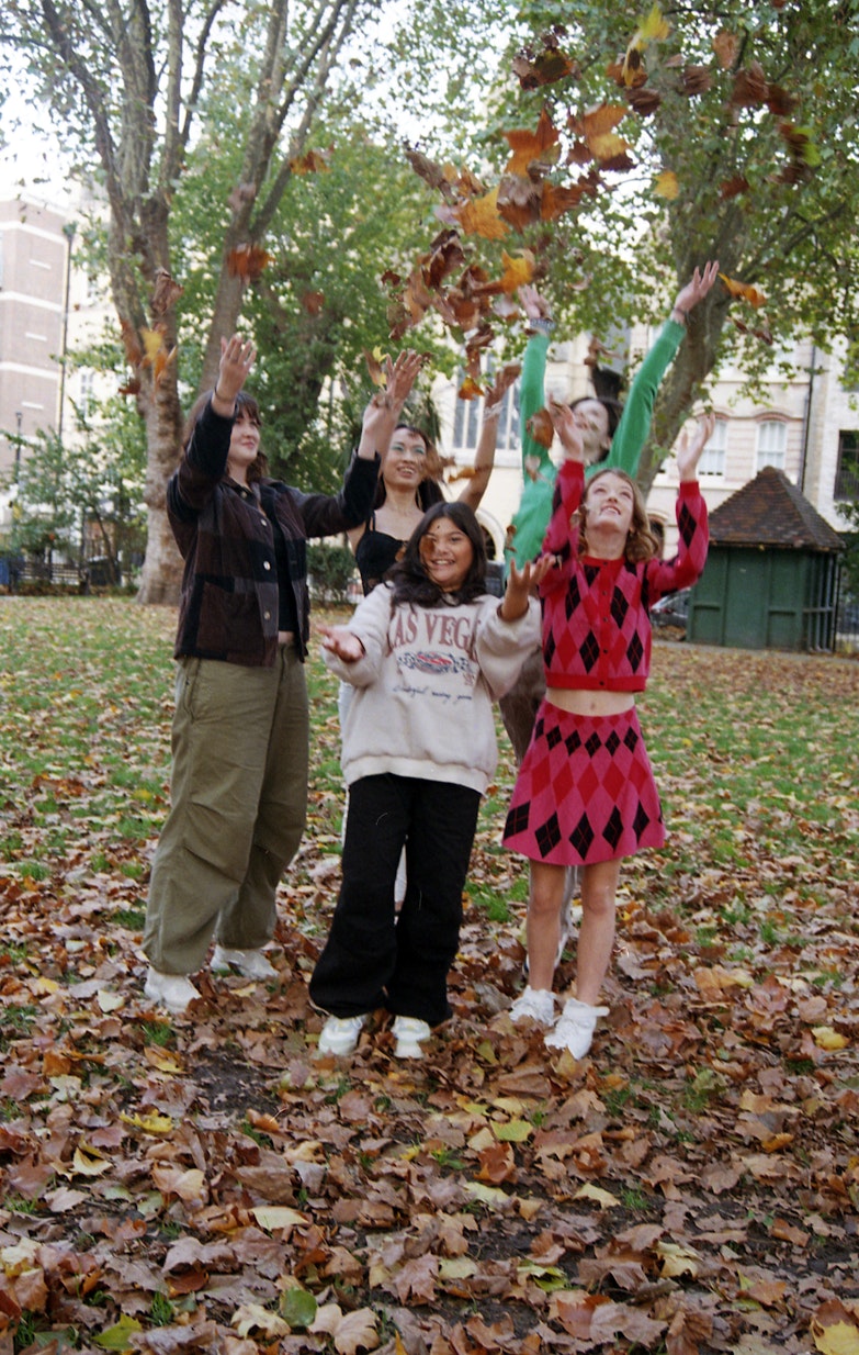 Four teenage girls outdoors in a park with autumn leaves on the ground, raising their arms and throwing leaves, one wearing a Las Vegas sweatshirt and another in a red diamond-pattern dress.