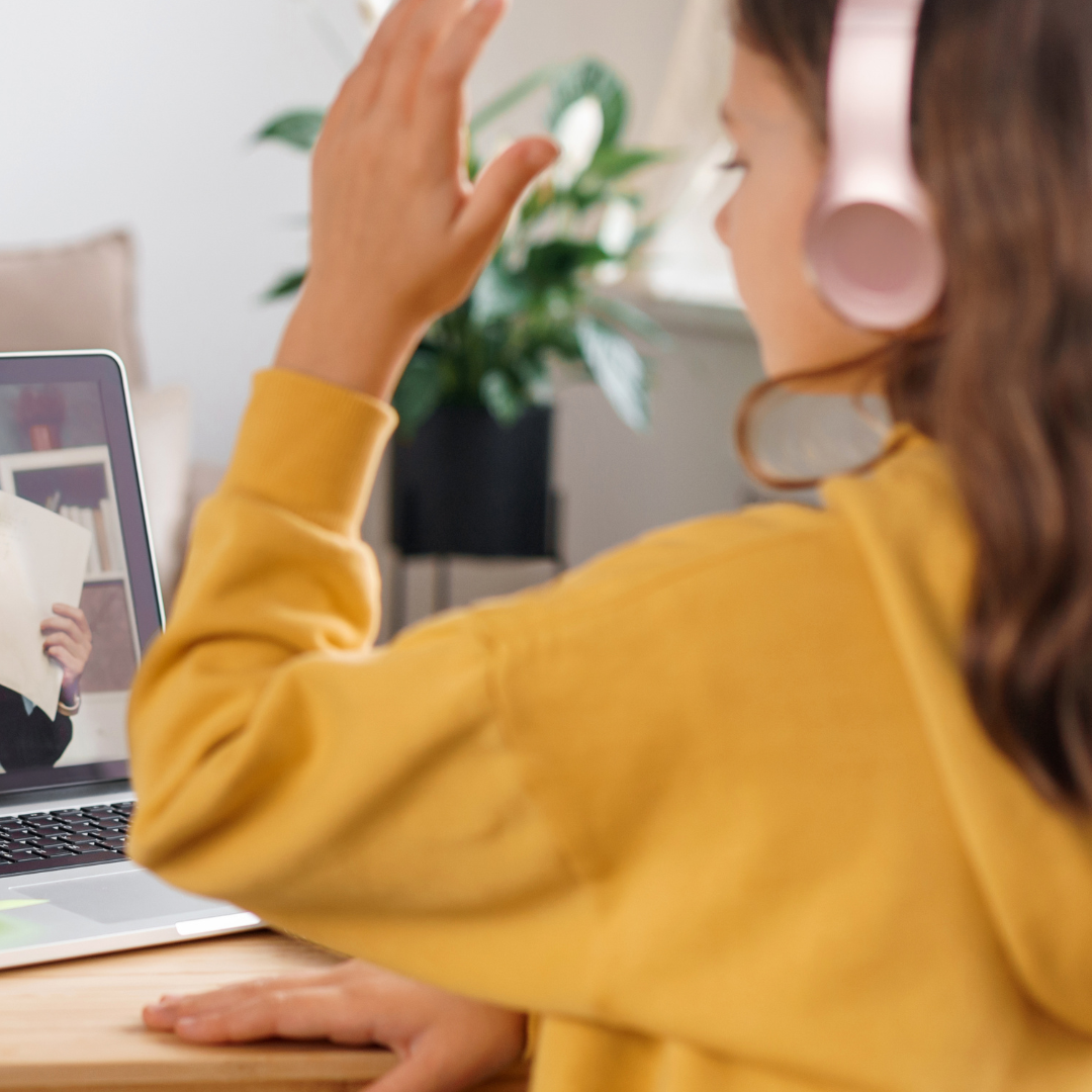 Teenage girl wearing pink headphones and a yellow jumper raises her hand while sitting at a desk in front of a laptop showing a video call with a tutor.
