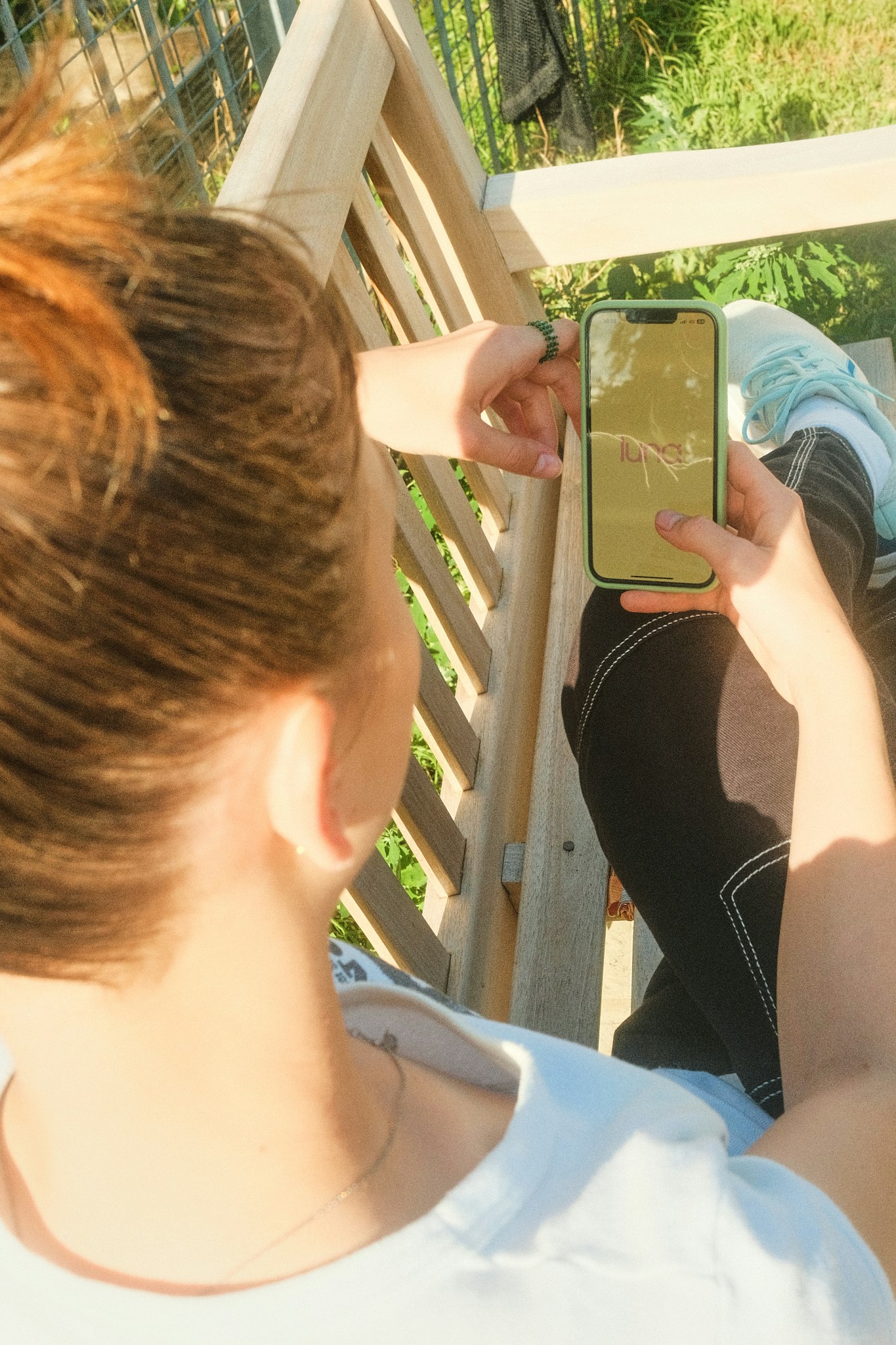 Close-up from above of a teenage girl with light brown hair holding up a phone showing the luna logo on screen, sitting outdoors on a wooden bench near greenery.