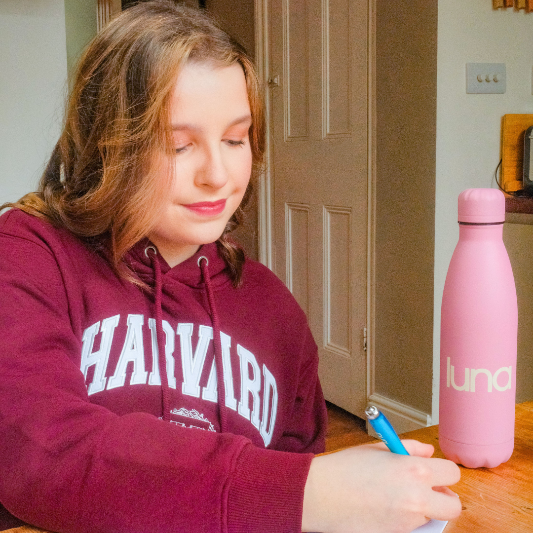 Teenage girl in a maroon Harvard hoodie sits at a table writing, with a pink luna water bottle beside her.