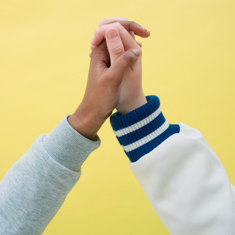 Two people clasping hands together against a plain yellow background, one wearing a grey sweatshirt and the other a white and blue striped-cuff top.