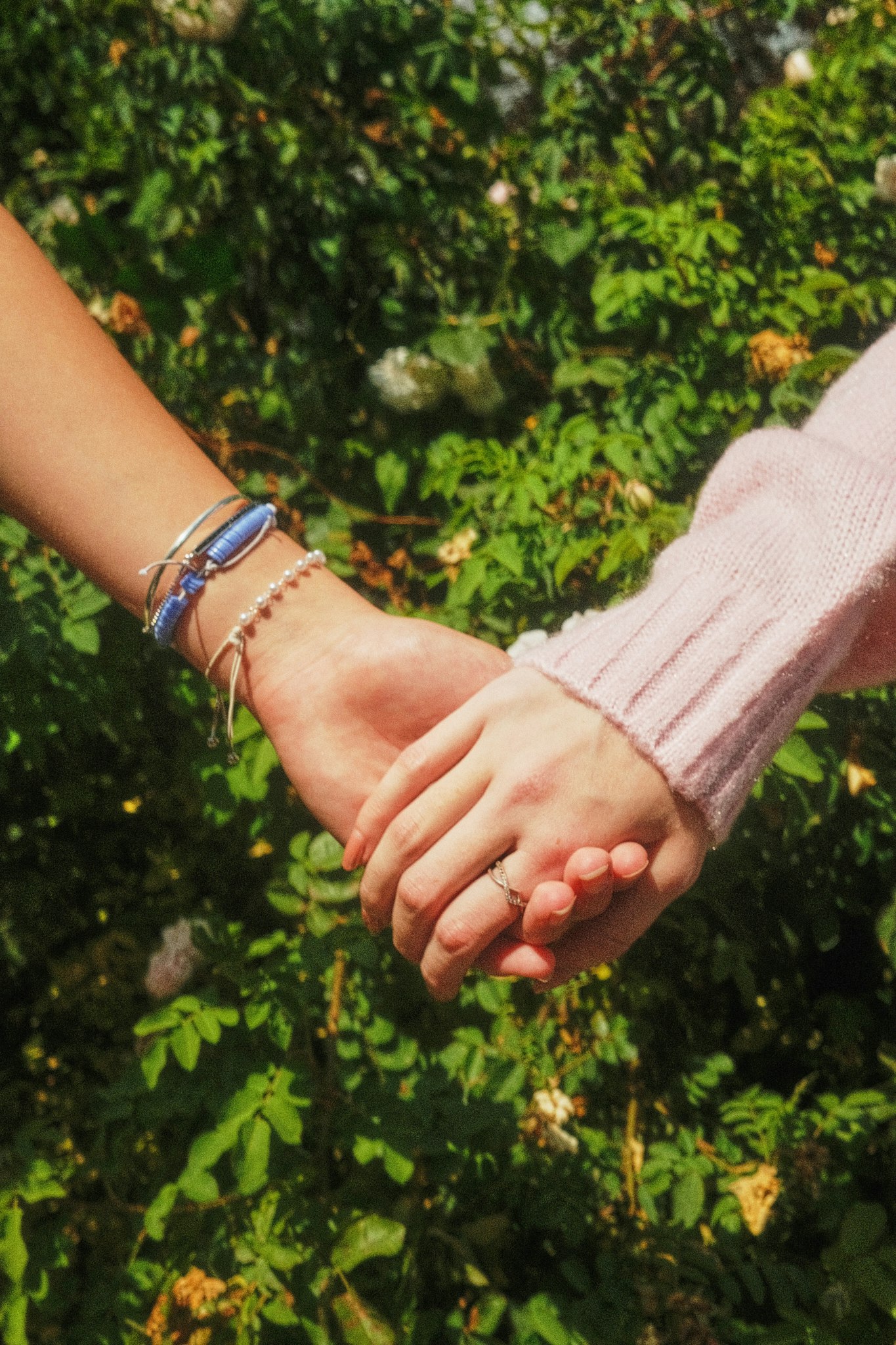 Close-up of two people holding hands outdoors against a leafy green background, one wearing beaded and blue bracelets and the other in a pink knitted sleeve.
