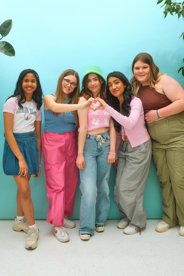 Five teenage girls of different body types standing together against a teal background, smiling and making heart shapes with their hands.