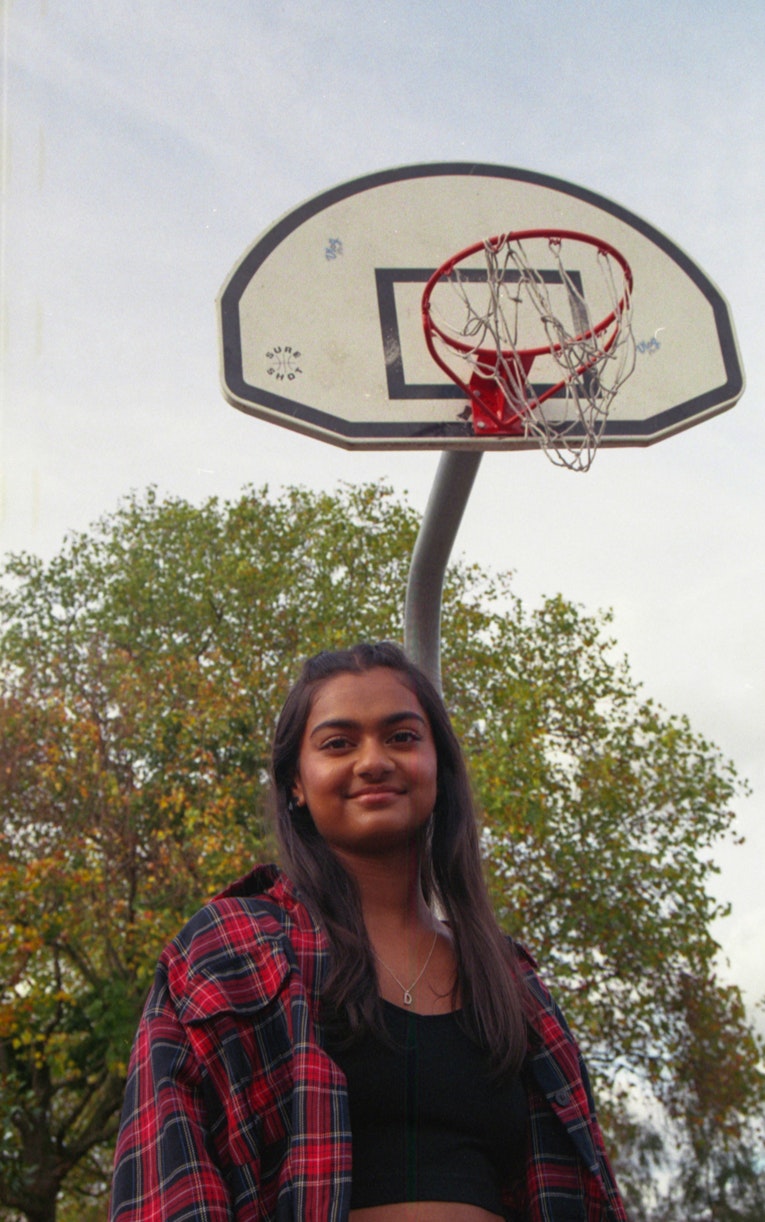 Teenage girl in a red tartan shirt smiling outdoors under a basketball hoop, with trees in the background.