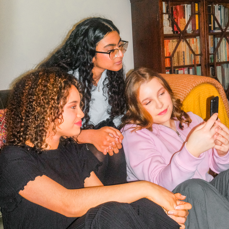 Three teenage girls sitting together on a sofa in front of a bookshelf, one holding a phone while the other two look on.