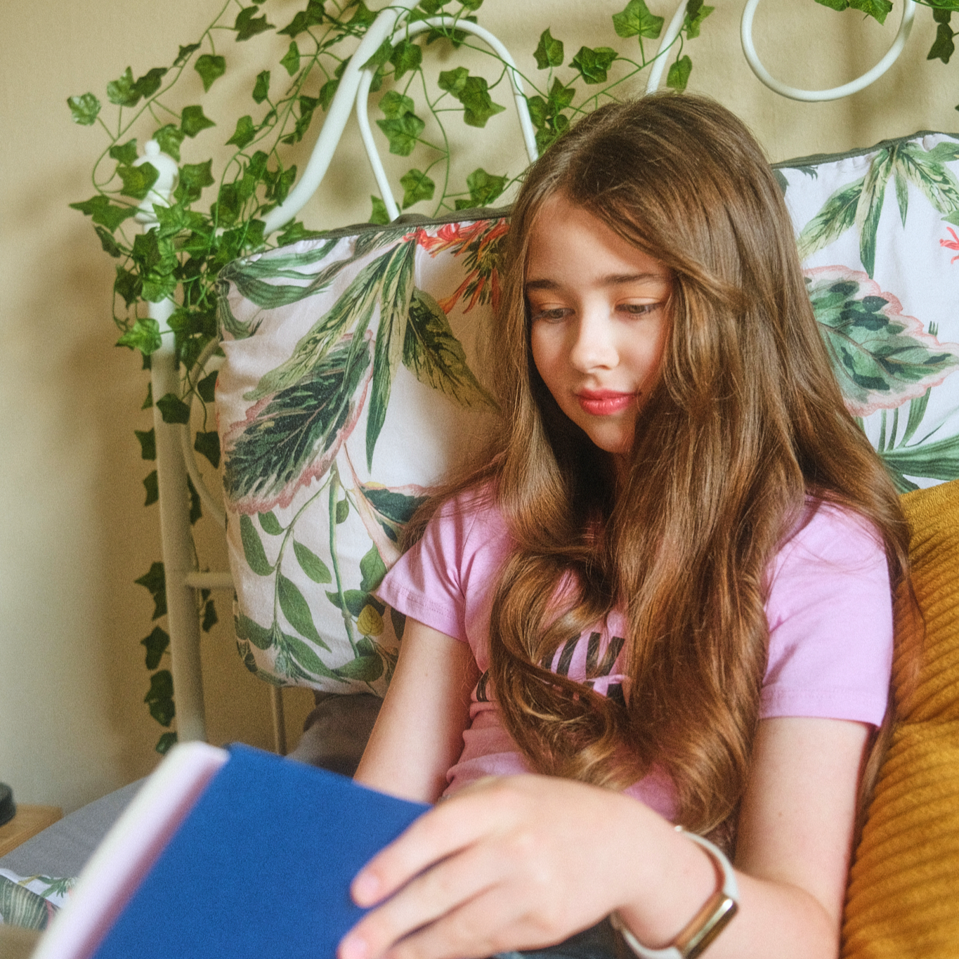 Teenage girl in a pink top sitting on a bed with leafy plant and patterned pillows behind her, holding a blue book.
