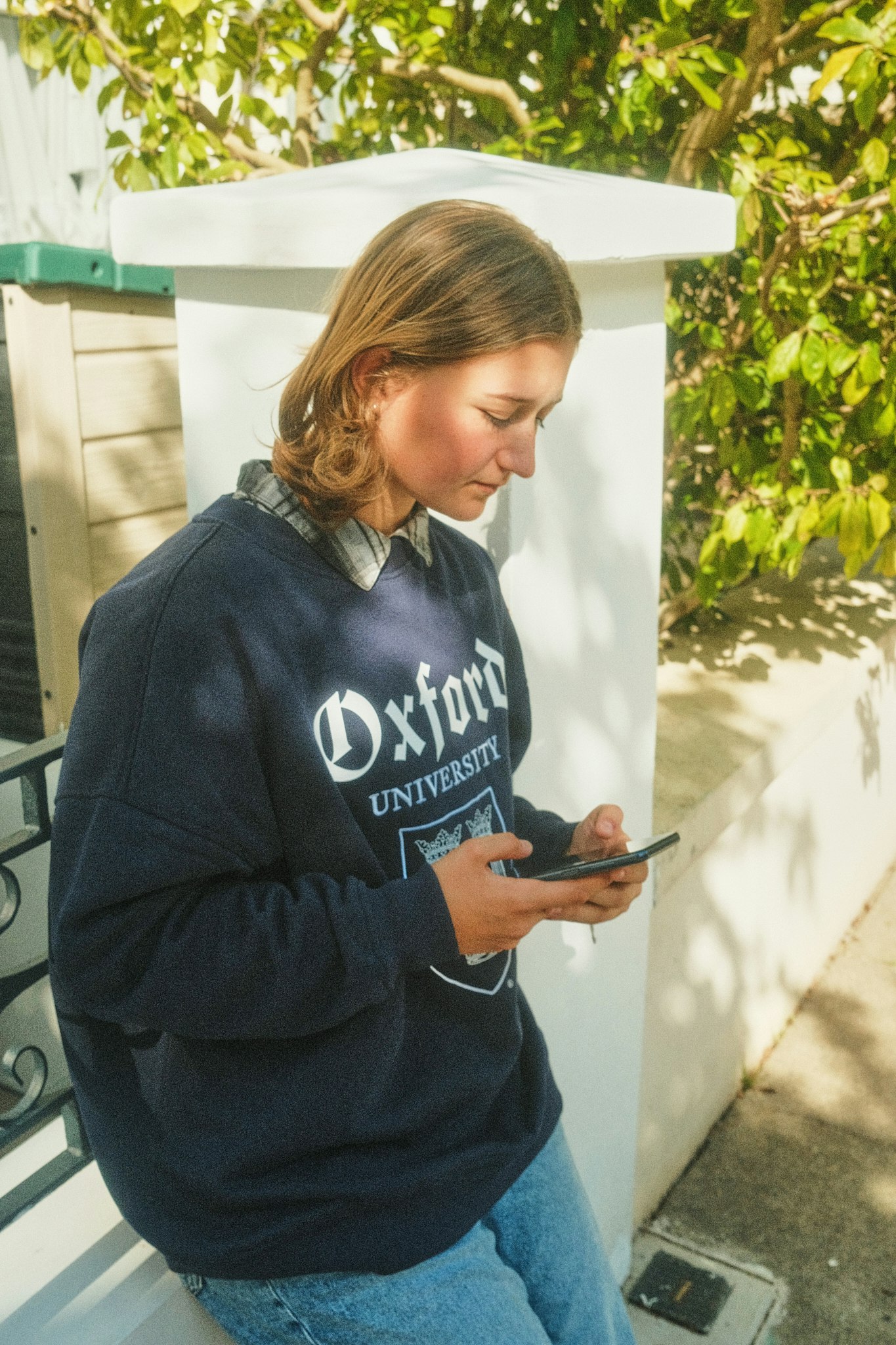 Teenage girl wearing a navy Oxford University hoodie standing outdoors looking down at her phone.