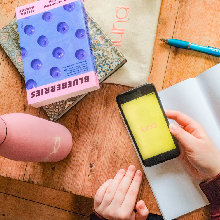 Overhead of a phone showing the luna app logo on a yellow screen, held by a hand, alongside a luna bag, a pink water bottle, books, an open journal, and a blue pen.