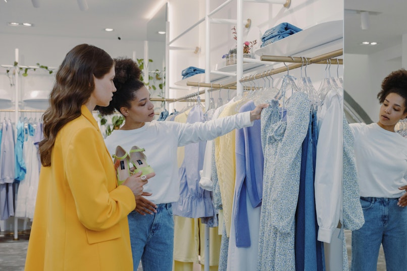 A woman in a yellow coat browses a clothes rail in a shop with another person holding sandals, in a bright store with plants and shelving.
