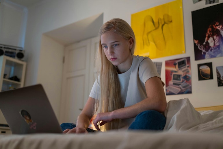 Teenage girl with long blonde hair sitting on a bed using a laptop at night, with posters and artwork on the wall behind her.
